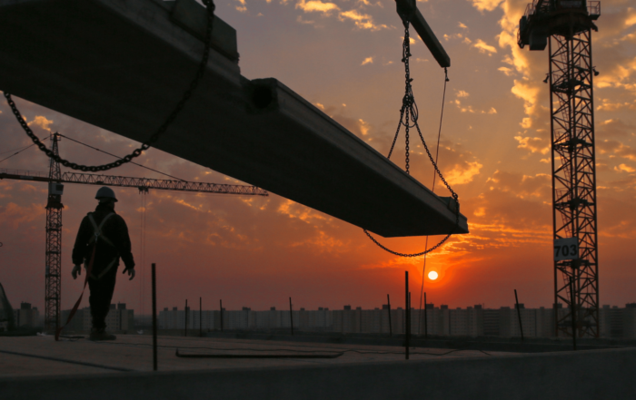 Construction worker looking off of the roof of a tall building towards an orange sunset. towering skyscraper in the background.