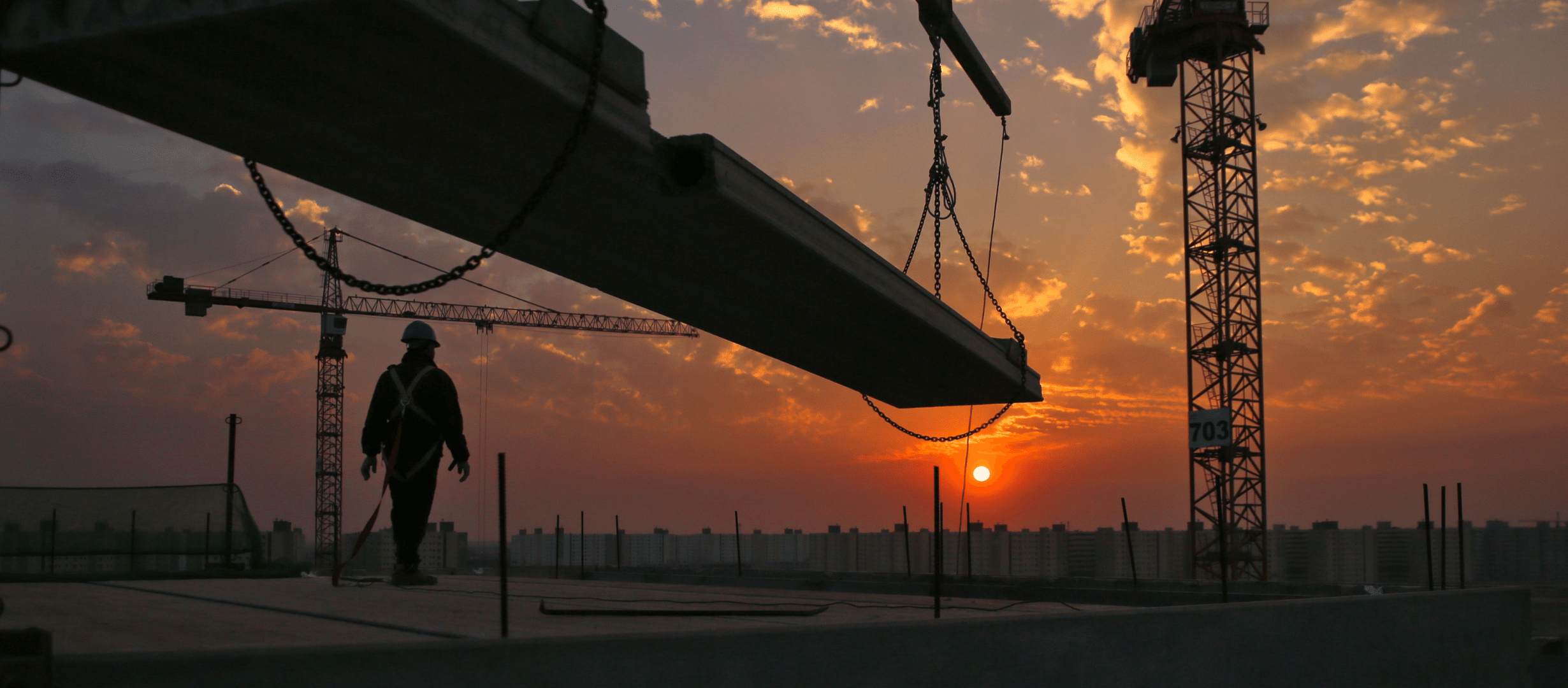 Construction worker looking off of the roof of a tall building towards an orange sunset. towering skyscraper in the background.